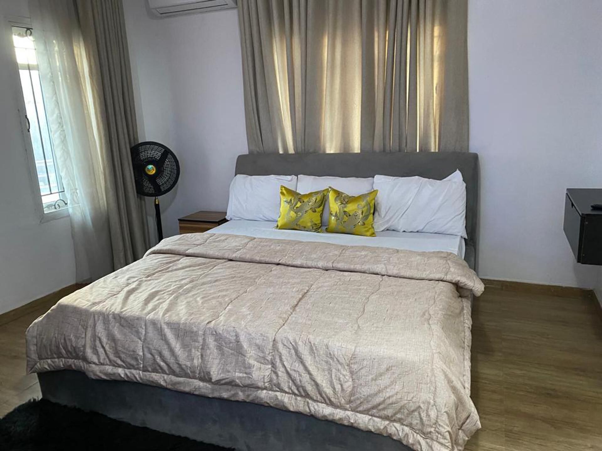 A neatly made bed with a grey headboard, white pillows, and a beige duvet. There is a black fan on a wooden nightstand beside the bed.