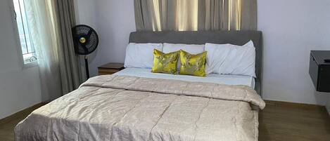 A neatly made bed with a grey headboard, white pillows, and a beige duvet. There is a black fan on a wooden nightstand beside the bed.
