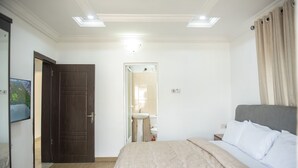 A bedroom with a bed, a nightstand, a mirror, a door, and a bathroom visible through an open door. - Captivating 3-bed Apartment in Lagos (Lekki)