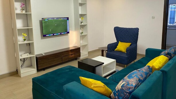 A modern living room with teal sofas, a blue armchair, and a flat-screen TV mounted on the wall. - Captivating 3-bed Apartment in Lagos (Lekki)
