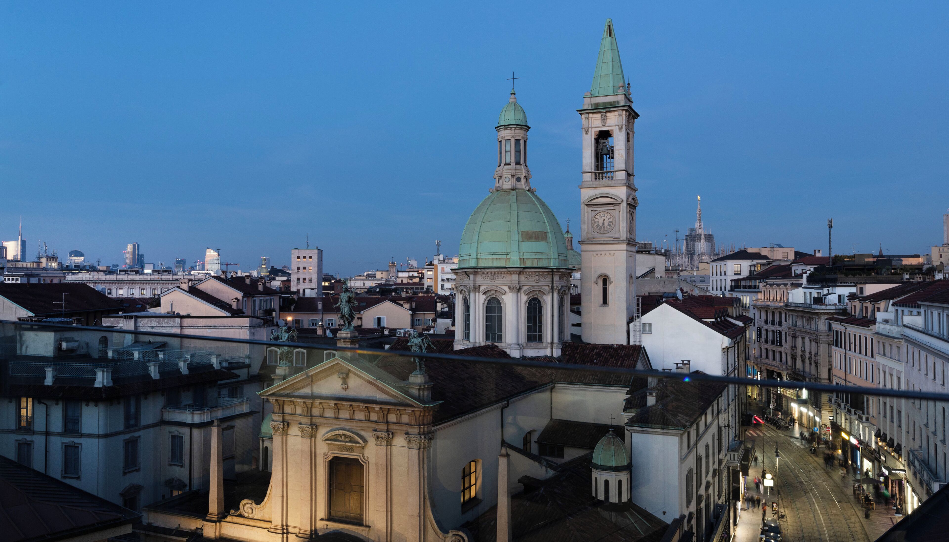 Paesaggio urbano con architettura storica, tra cui una chiesa in evidenza con una cupola verde e un'alta torre dell'orologio.