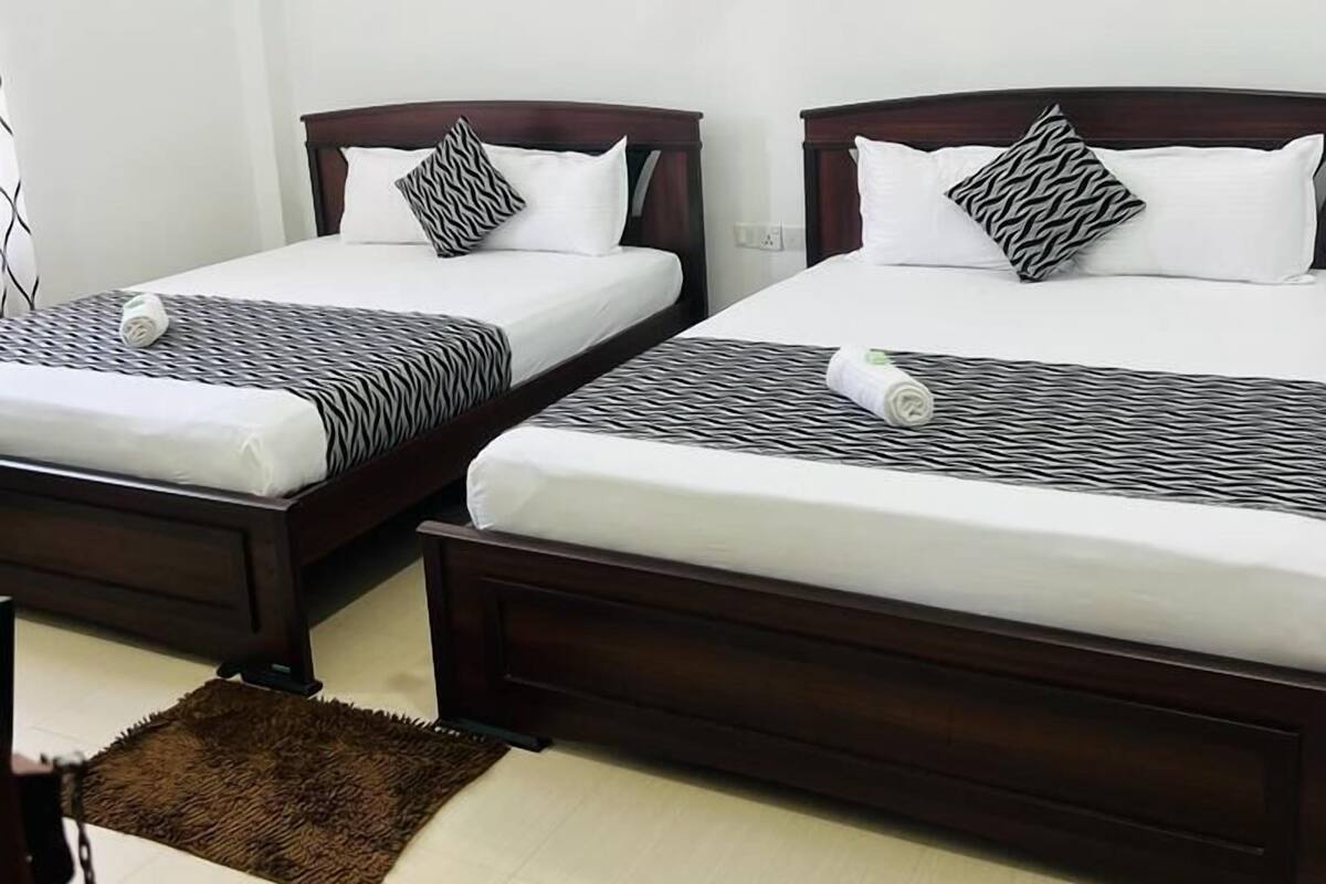 Une chambre d’hôtel avec deux lits en bois, du linge de lit blanc, des coussins à motifs zébrés, un tapis marron, un ventilateur de plafond et un tableau représentant un paysage accroché au mur.