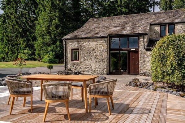Een gebouw van steen met een houten terras, een tafel en vier rieten stoelen.
