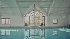 An indoor swimming pool with clear glass walls, two lounge chairs, and a view of trees outside. - Meadow 2 Bedroom Lodge 1 (Umberleigh)