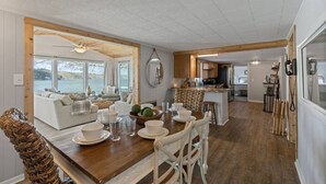 Een ruime woonkamer met een houten eettafel voor vier personen, een gezellige zithoek met uitzicht op een meer, en een keuken op de achtergrond.