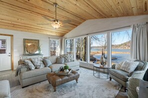 Ein gemütliches Wohnzimmer mit Holzbalkendecke, großen Fenstern mit Seeblick, einem Lederhocker und einer Couch mit gemusterten Kissen.