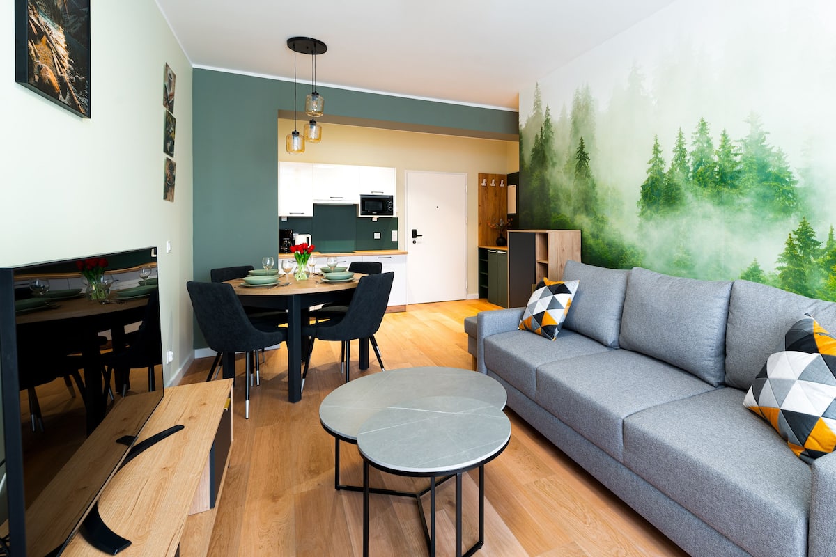 A modern living room with a grey sofa, a dining table with chairs, and a kitchen in the background.
