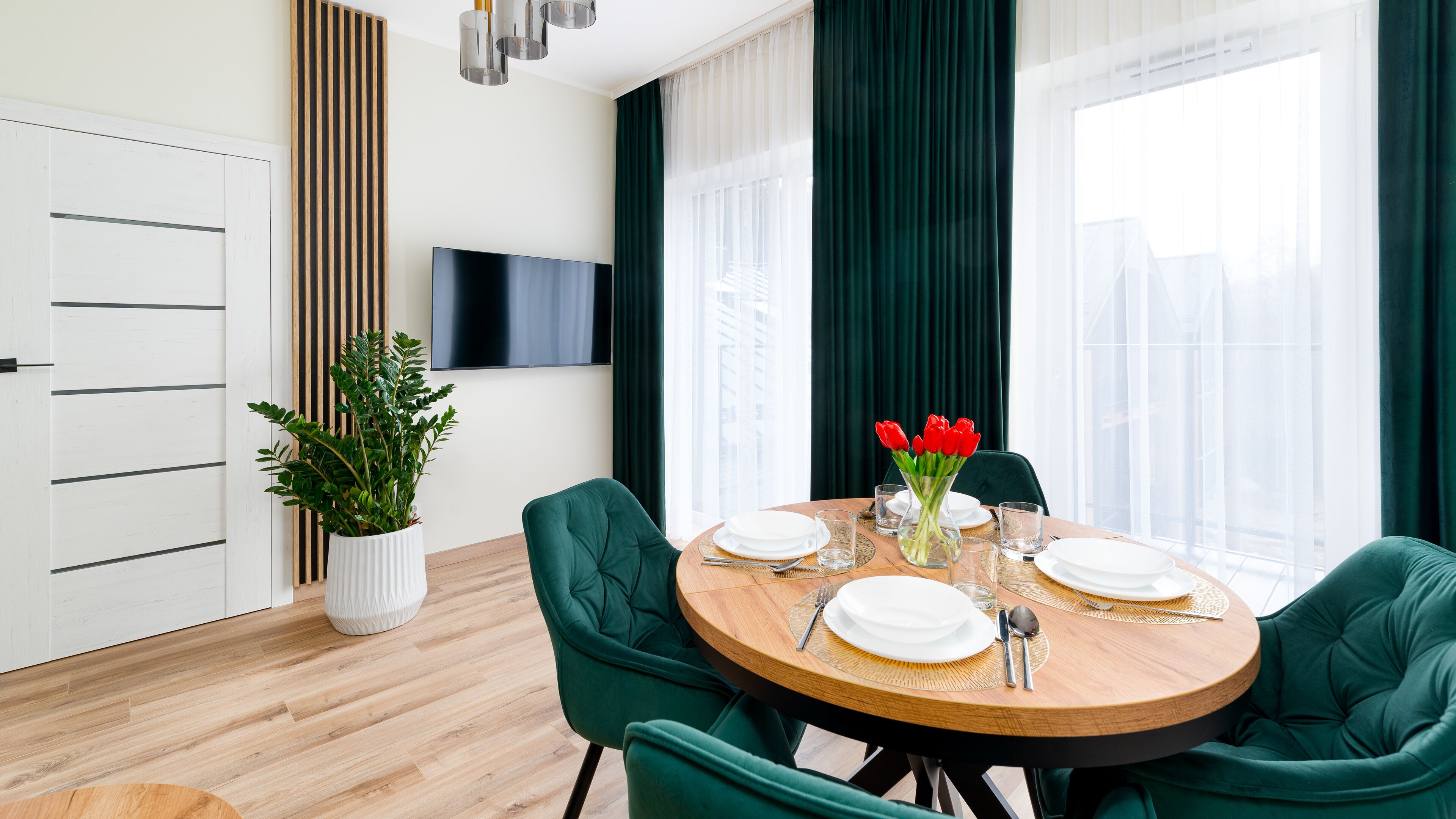 A modern dining area with a round wooden table set for four, surrounded by green upholstered chairs. A potted plant and a vase with red flowers are on the table.