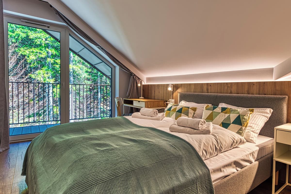 A modern bedroom with a large bed, a nightstand, a wooden headboard, and a view of a forest through a balcony.
