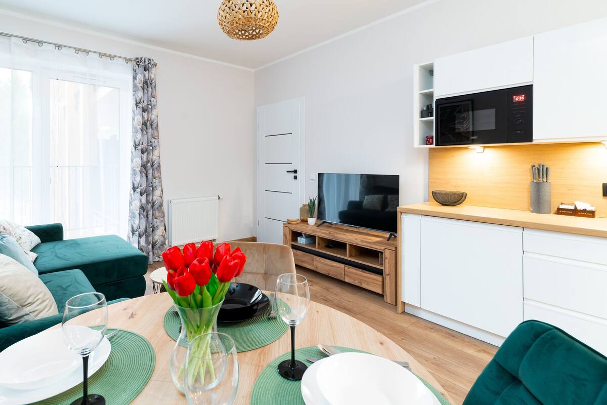 A modern living room with a green sofa, a dining table set with plates and wine glasses, and a TV in the background.