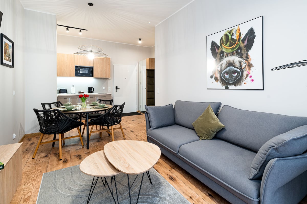 A modern living room with a grey sofa, a dining table with chairs, and a kitchen in the background.