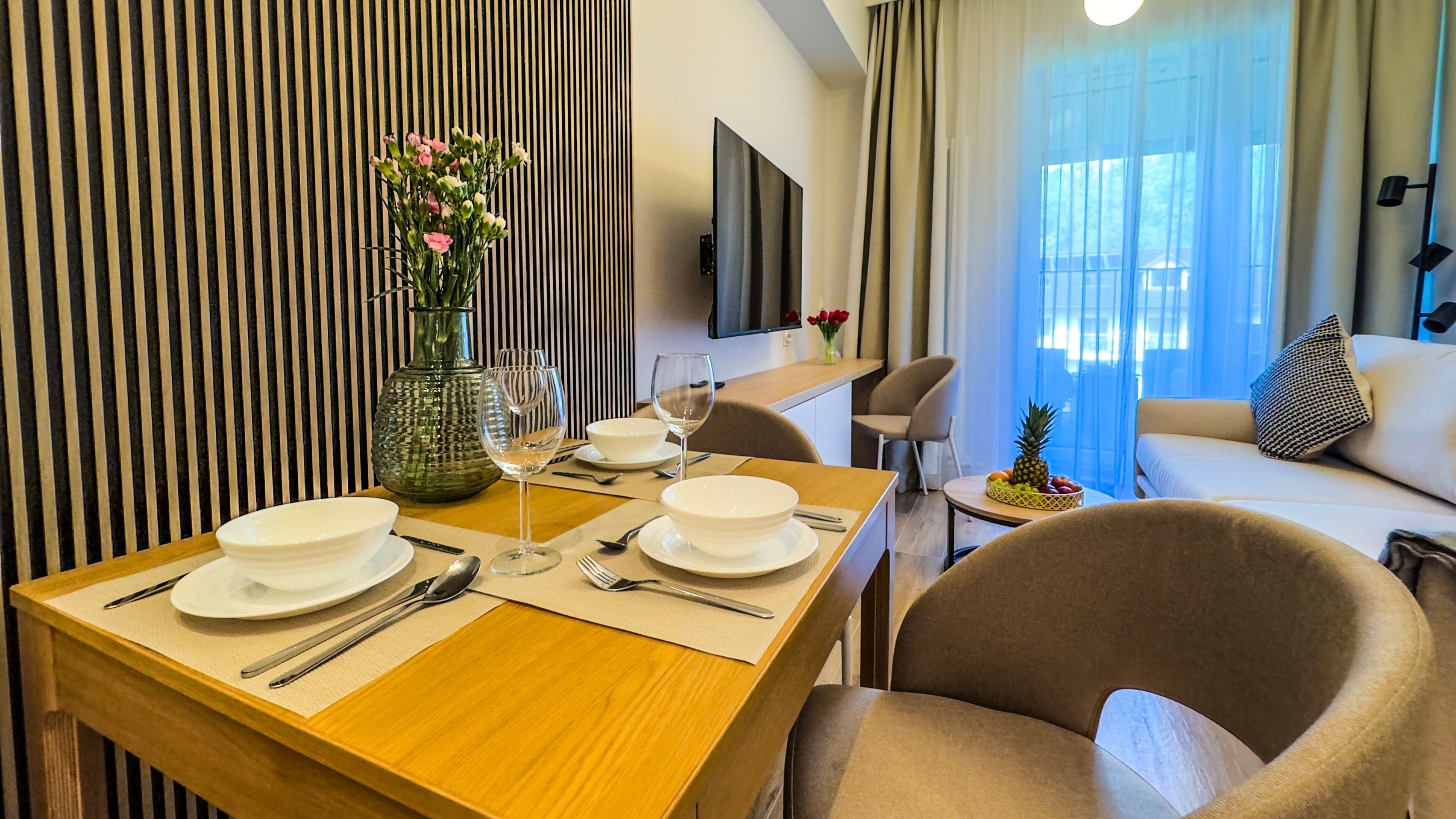 A dining area with a wooden table set for two, a vase with flowers, and a television mounted on the wall.