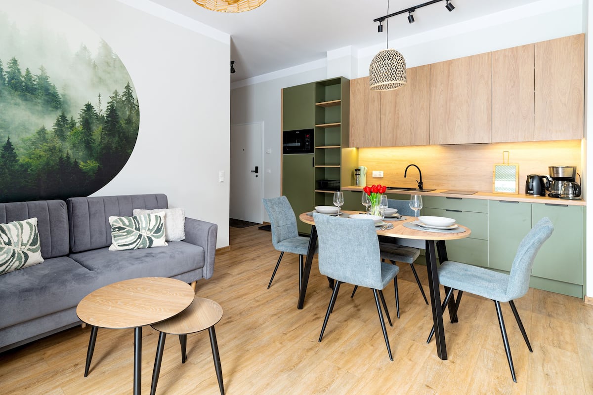 A modern living room with a grey sofa, round wooden tables, and grey chairs. A large circular painting of a forest is on the wall.
