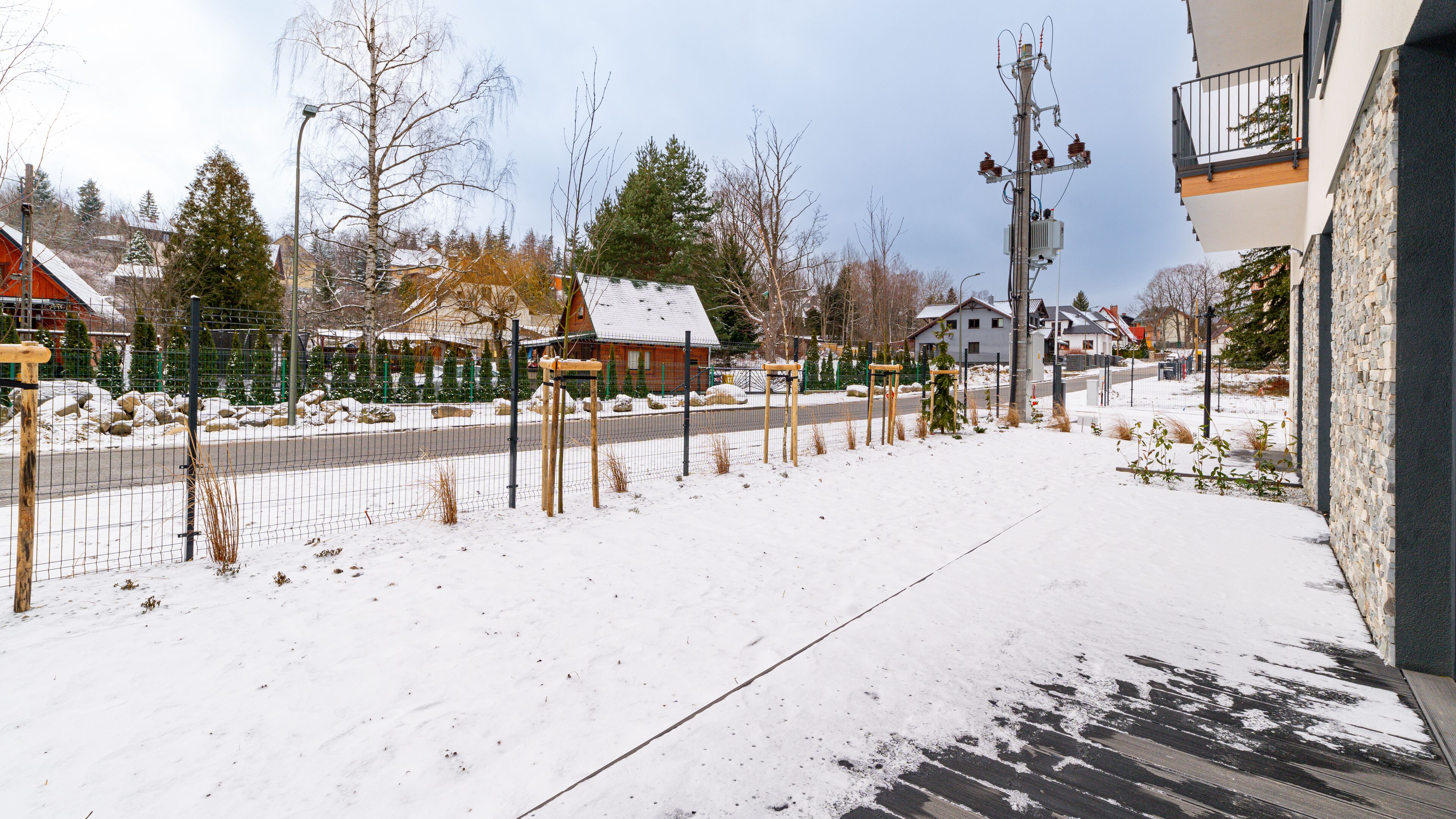 A snow-covered outdoor area with a fence, a wooden deck, and houses in the background.