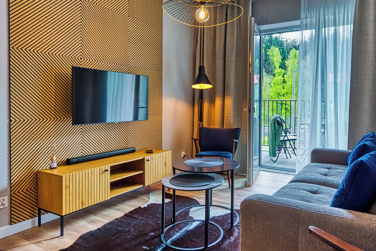 A modern living room with a flat-screen TV, wooden panel wall, a sofa with blue cushions, a small round table, and a chair.