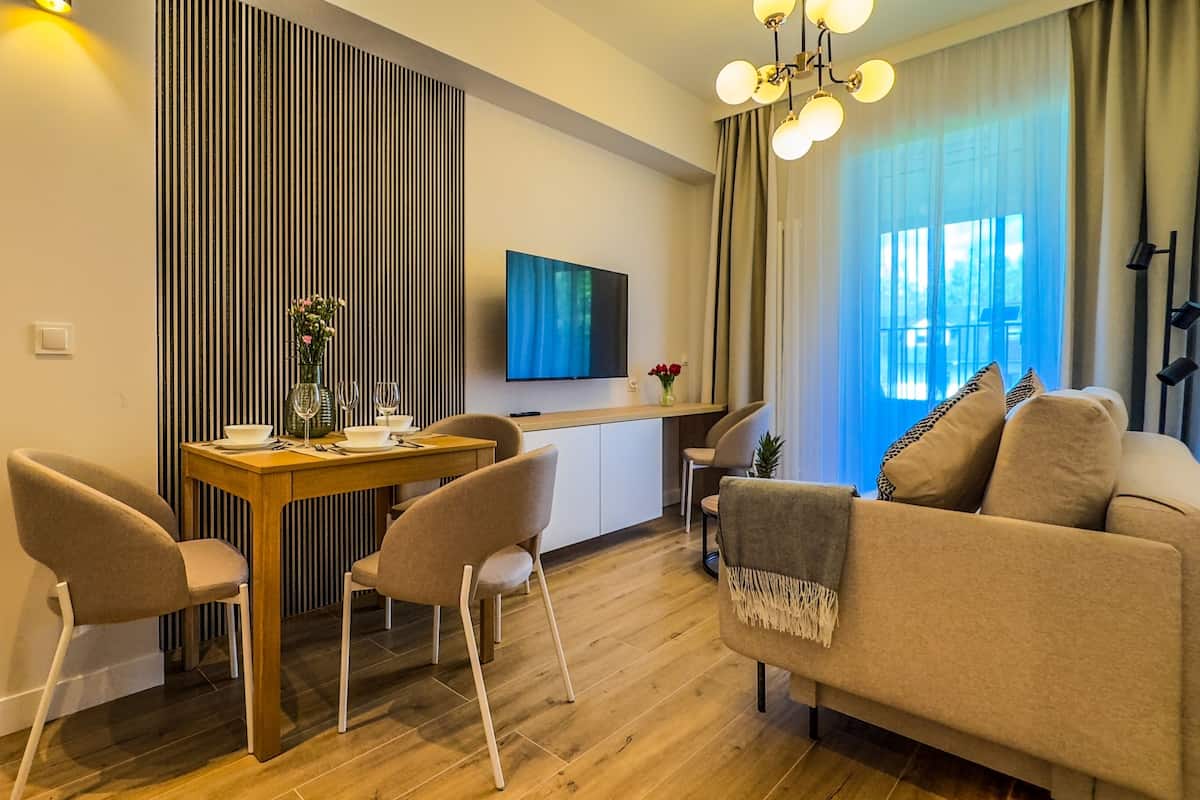 A modern living room with a sofa, dining table, and chairs. A flat-screen TV is mounted on the wall, and there is a window with curtains.