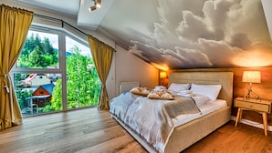 A bedroom with a large bed, a nightstand, a lamp, and a view of a forest through a sliding door.