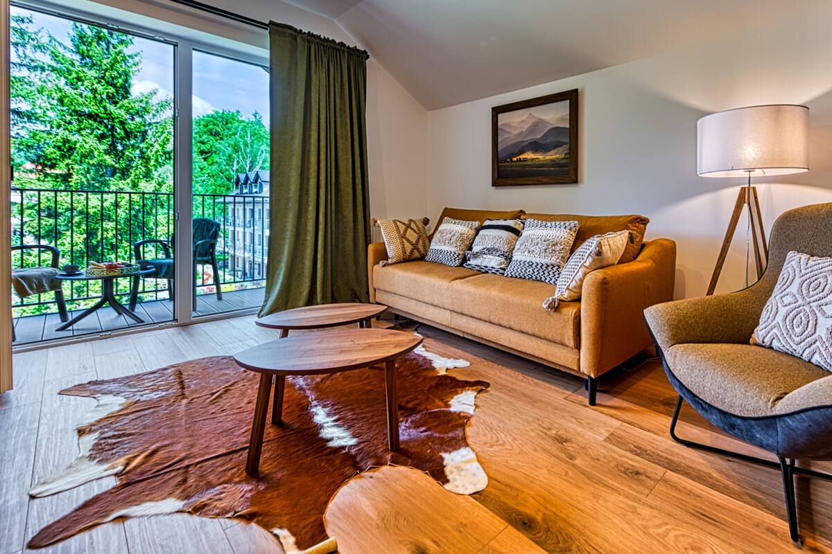 A modern living room with a sofa, armchair, and floor lamp. There is a large window with a view of greenery and a balcony.