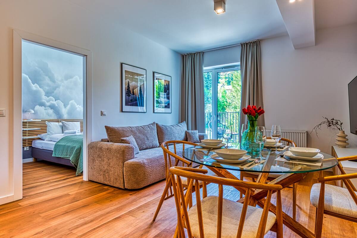 A modern living room with a glass dining table, wooden chairs, a sofa, and a bed with a green blanket.
