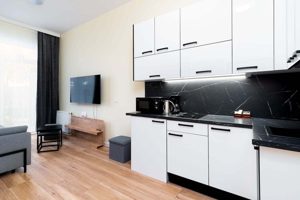 A modern kitchen with white cabinets, a black countertop, and a black marble backsplash. There is a grey sofa and a small table in the living area.