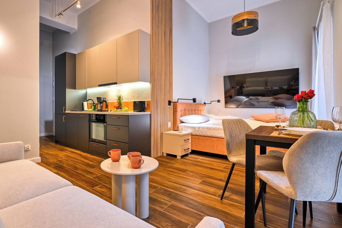 A modern hotel room with a kitchenette, a bed with a headboard, a dining table with chairs, and a small round table with two cups.