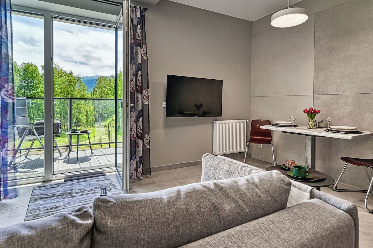 A modern living room with a large sliding glass door leading to a balcony, a flat-screen TV mounted on the wall, a dining area with a table and chairs, and a sofa.