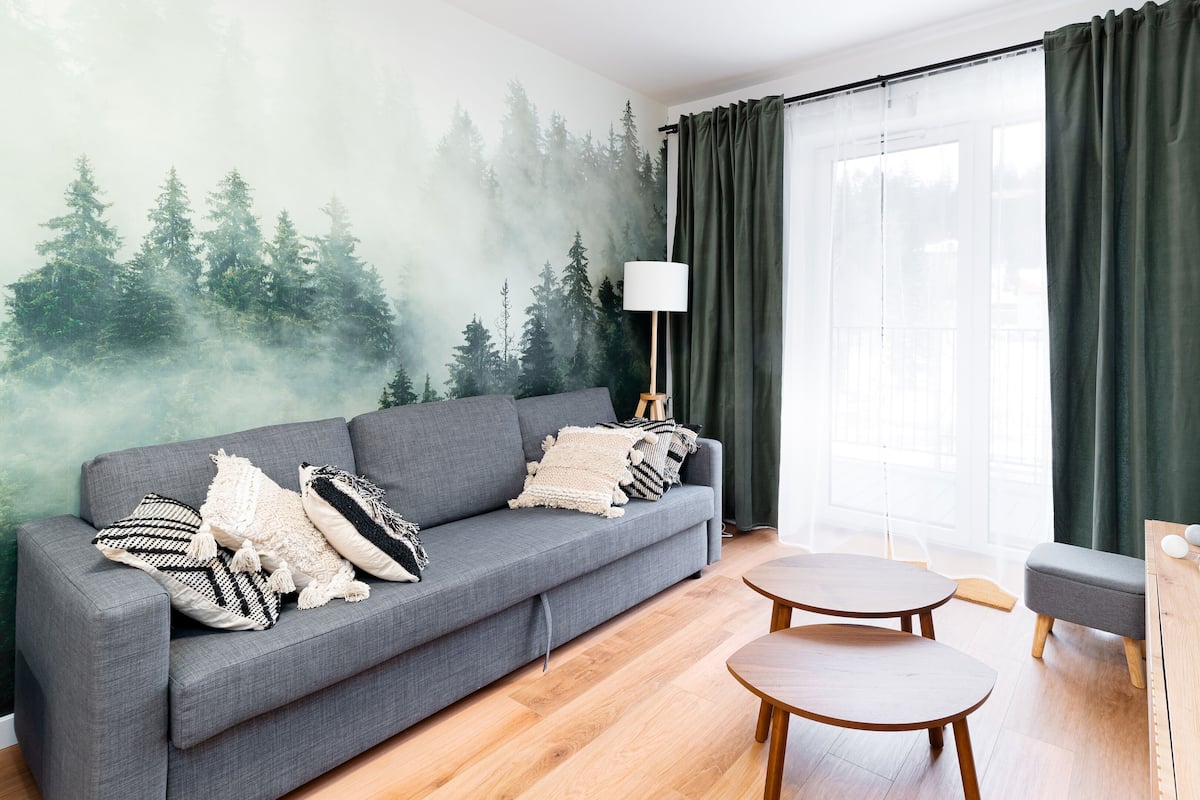 A modern living room with a grey sofa, two wooden side tables, a lamp, and a wall mural of a forest.