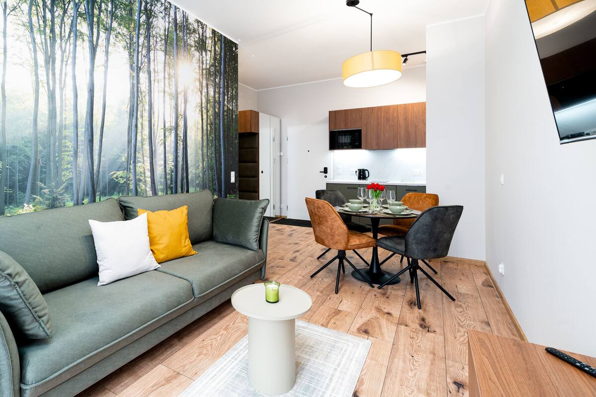 A modern living room with a green sofa, a dining table with chairs, and a kitchen area in the background.