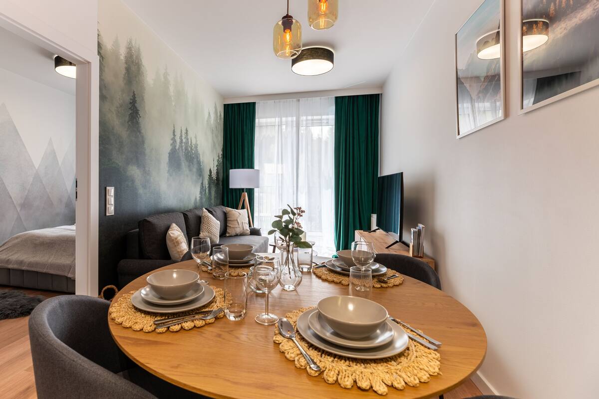 A dining area with a round table set for four, a sofa, and a painting of a forest on the wall.
