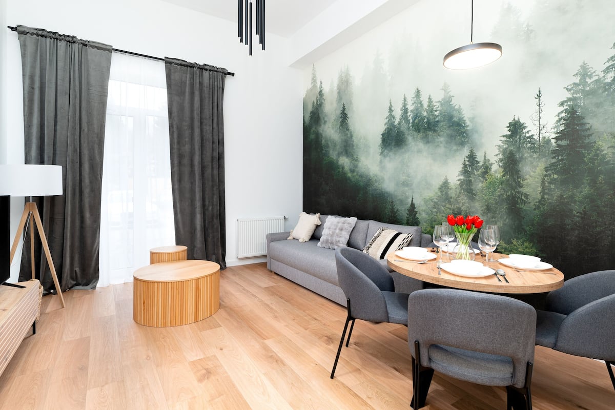 A modern living room with a sofa, dining table, and chairs. There is a painting of a forest on the wall.