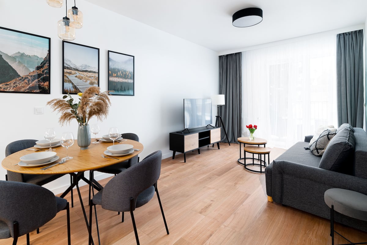 A modern living room with a dining area, a sofa, and a TV. There are three framed pictures on the wall, a vase with dried flowers, and a lamp.