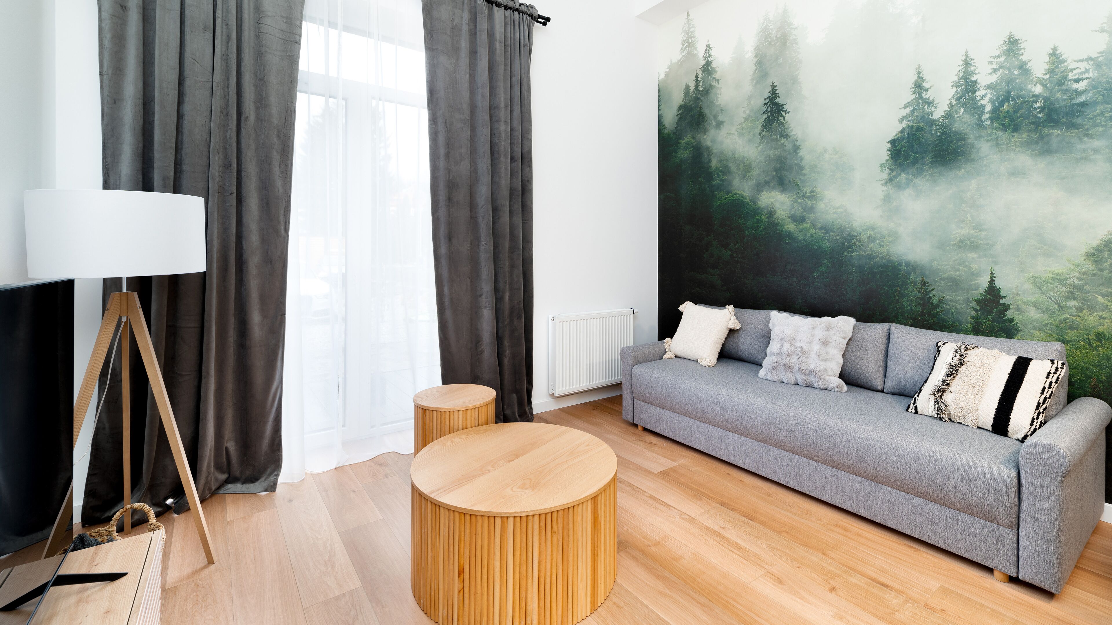 Un salon moderne avec un canapé gris, des petites tables d’appoint rondes en bois, un lampadaire et une fresque représentant une forêt sur le mur.