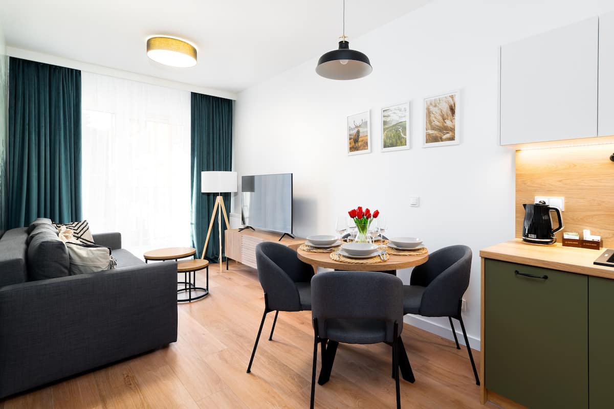 A modern living room with a sofa, coffee table, and dining area with chairs and a table set for two. A TV and floor lamp are visible in the background.