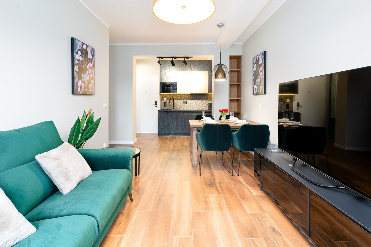 A modern living room with a teal sofa, a flat-screen TV, and a dining area with a wooden table and chairs.