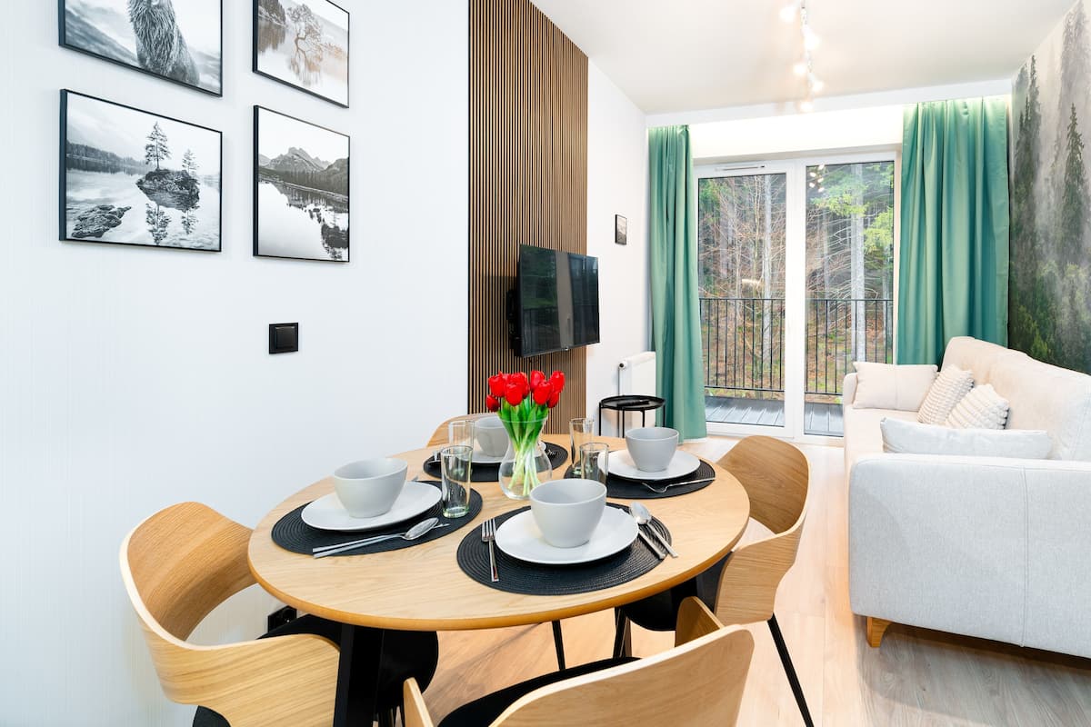 A modern living room with a sofa, dining table, and chairs. There are framed pictures on the wall and a large window with curtains.