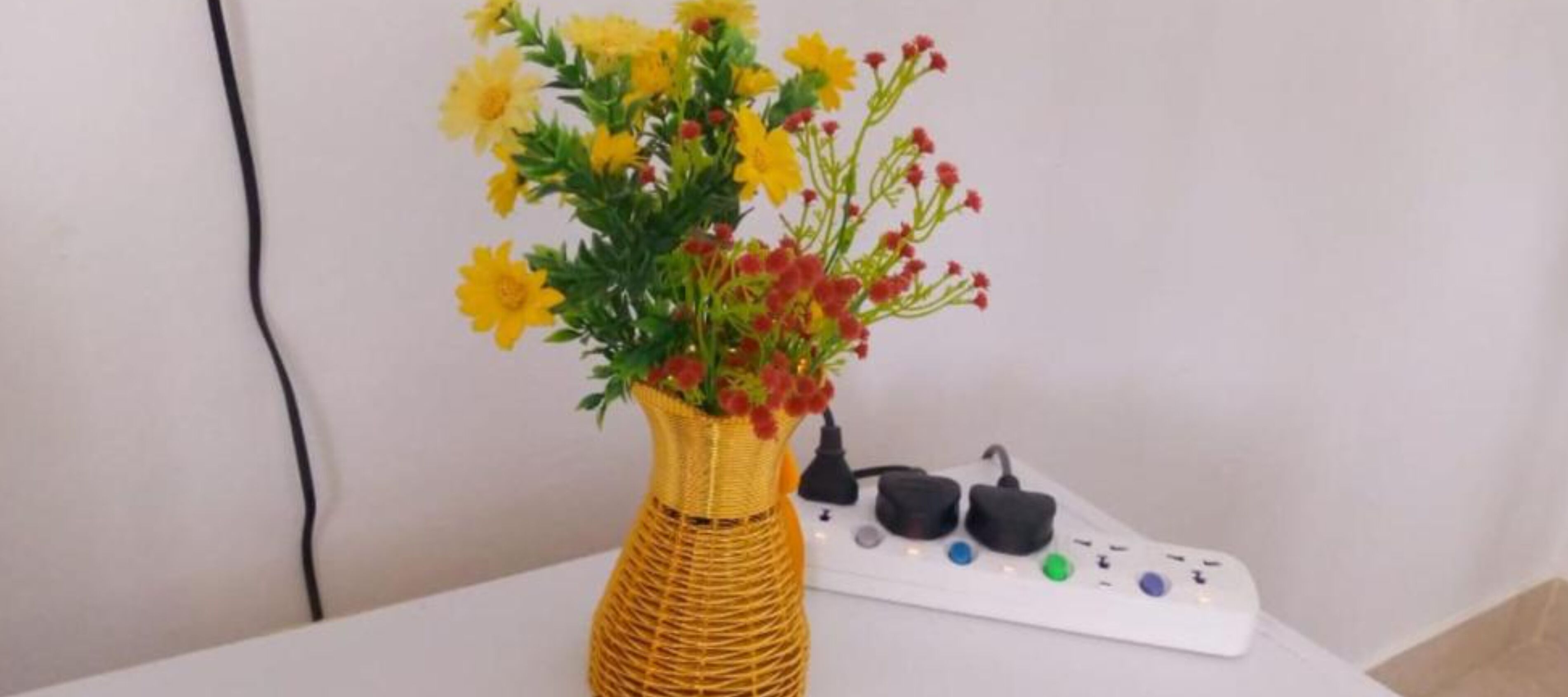 Un florero de mimbre con flores amarillas y rojas sobre una mesa blanca, junto a un enchufe blanco y un control remoto.