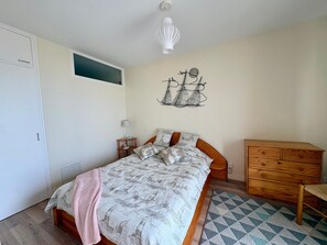 Une chambre à coucher avec un lit, une commode en bois, un luminaire fixé au mur et une petite fenêtre.
