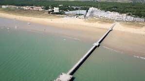 Eine Luftaufnahme einer Küstenregion mit einer langen Mole, die sich ins Meer erstreckt, einem Sandstrand und einer Ansammlung von Gebäuden im Hintergrund.