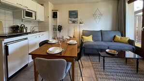 A modern kitchen with white cabinets, a microwave, and a sink. There is a dining table set with plates, glasses, and cutlery. A grey sofa and a small table with a lamp are in the living area. - Cozy Apartment in Montgenevre - Sleeps 4 (Montgenèvre)