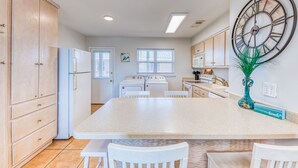 A kitchen with wooden cabinets, a large island, white chairs, and a clock on the wall. - 2BR Beach Getaway w/ Pool walkable to The Hangout!  ~ Walk to beach in 3 minutes (Gulf Shores)