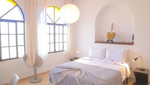 A bedroom with a bed, two bedside lamps, a chair, a large window with curtains, and a wall-mounted shelf with decorative items. - Villa Roka : Magnifique Villa Avec Piscine, Dans un Endroit Calme et Reposant (Essaouira)