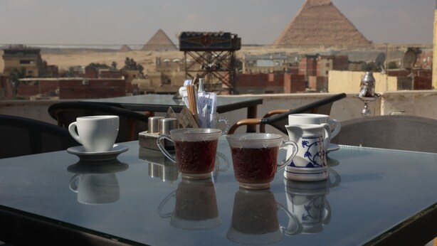 A table with two cups and a teapot, set against a backdrop of pyramids and a desert landscape. - Eagles pyramids view - Foriigners only (Giza)