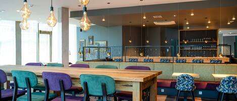 A modern restaurant with a long wooden table, colorful chairs, and hanging light fixtures.