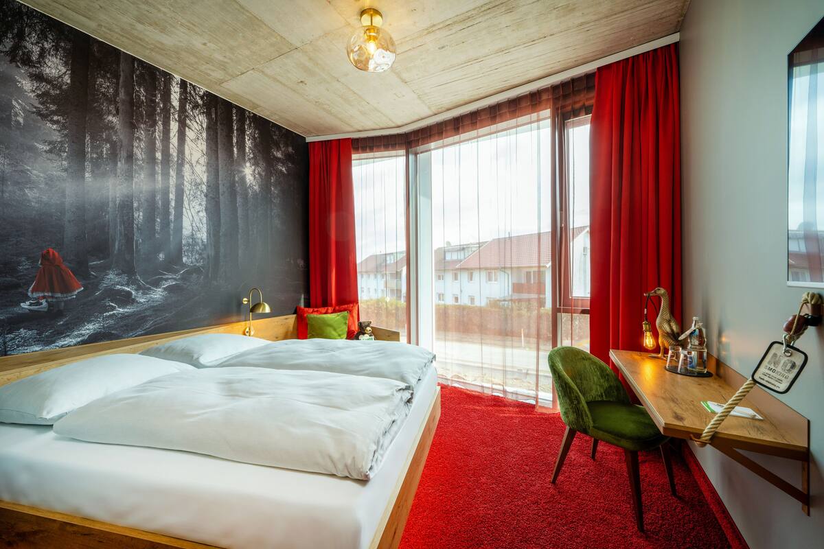 Un dormitorio con una cama grande, un escritorio, una silla, una alfombra roja y un mural con un bosque en la pared.
