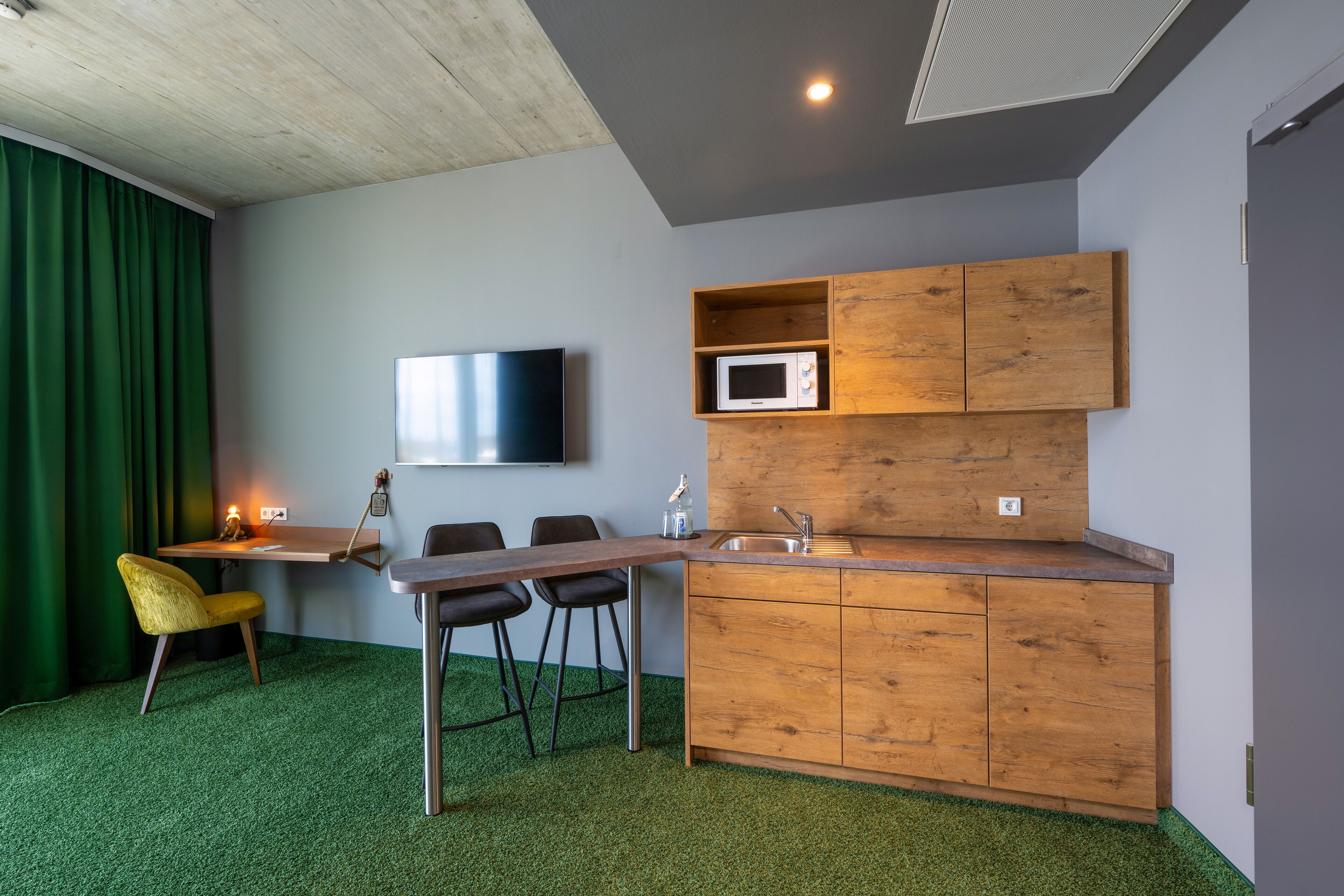 A compact kitchen with a wooden countertop, a microwave, and a sink. There is a bar stool and a chair, and a flat-screen TV mounted on the wall.