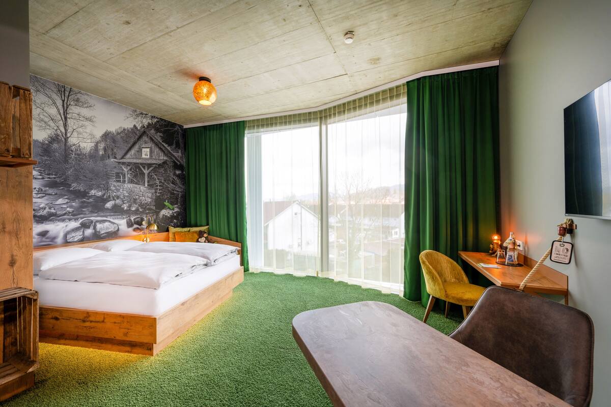 Una habitación con una cama, un escritorio de madera, una silla y un ventanal con cortinas.