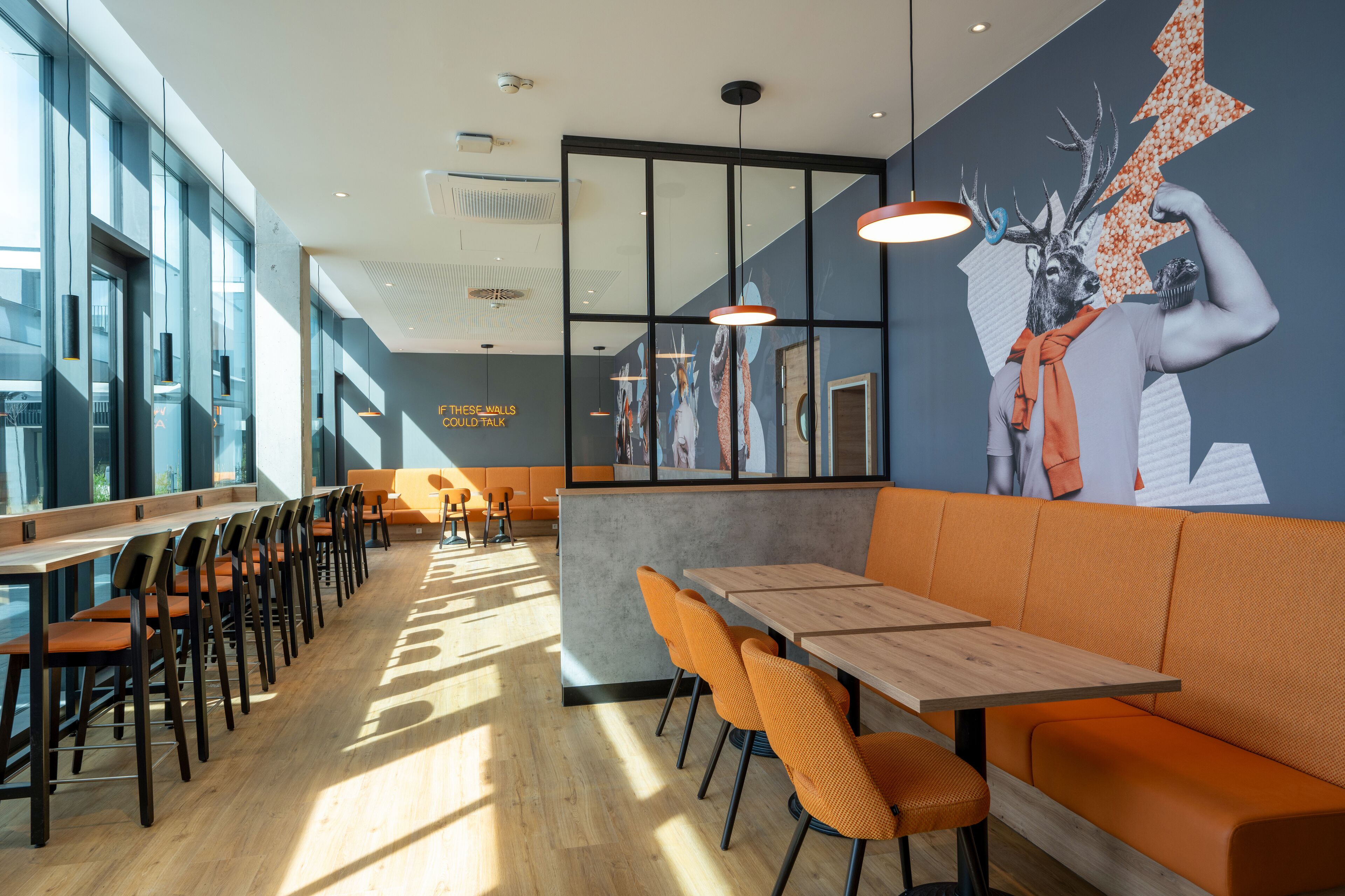 Un restaurante moderno con mesas de madera largas y asientos acolchados color naranja, ventanas amplias y un mural en la pared.