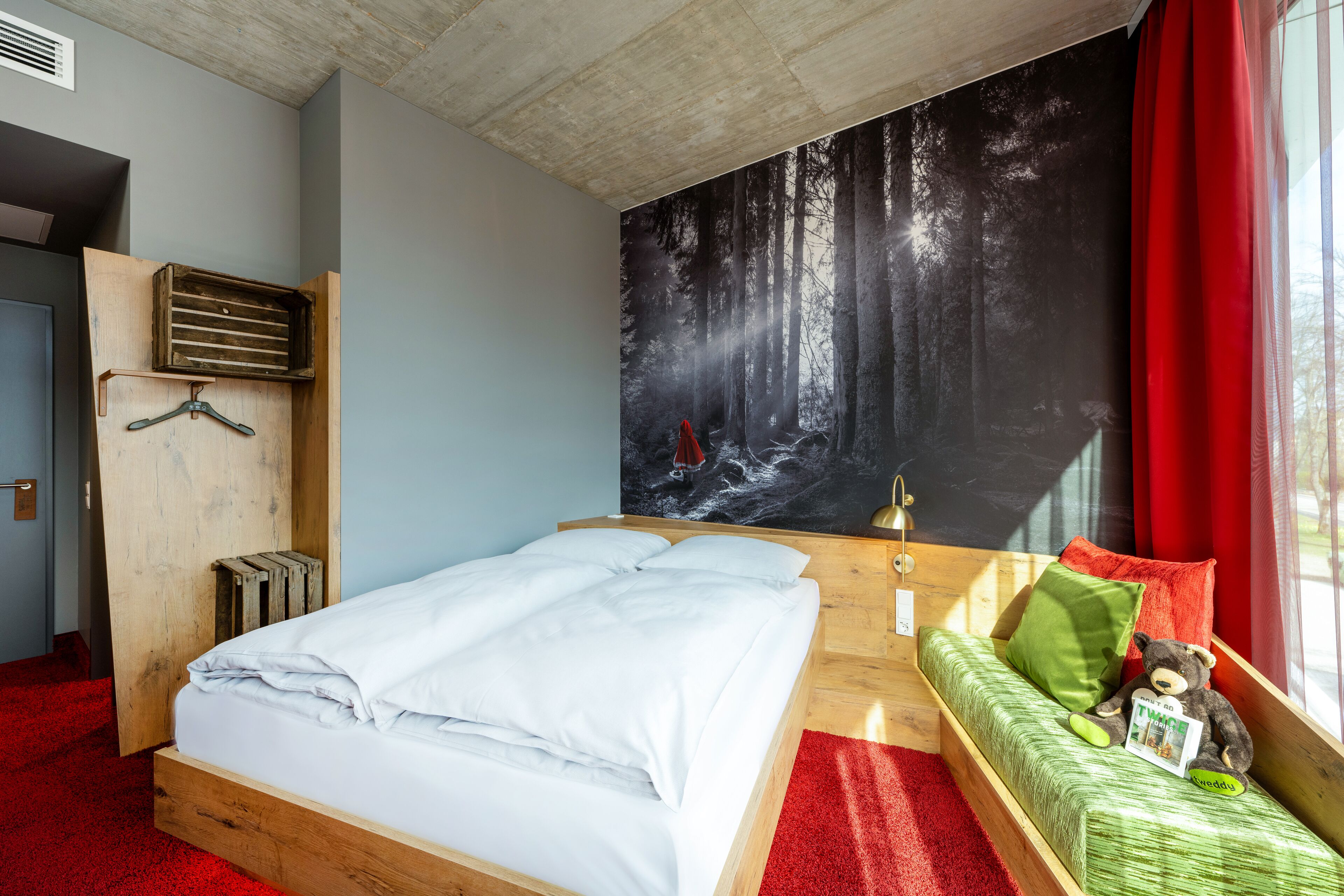A bedroom with a bed, a wooden bench with a teddy bear, a red curtain, and a forest mural on the wall.