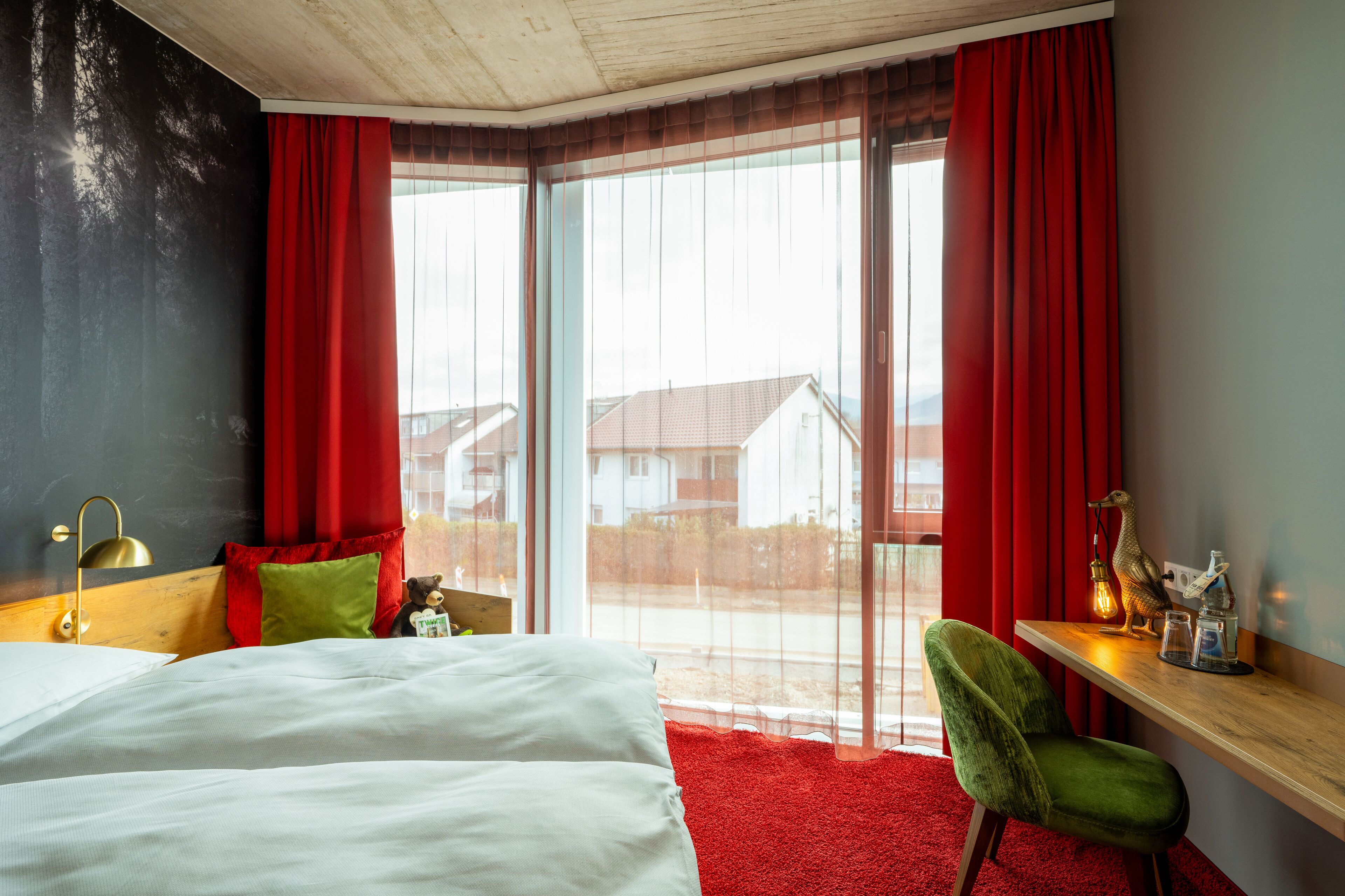 A hotel room with a bed, a green chair, a wooden desk, and a large window with red curtains.
