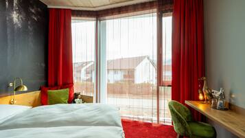 A hotel room with a bed, a green chair, a wooden desk, and a large window with red curtains.